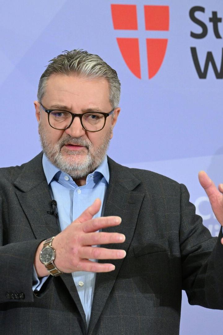 Peter Hacker steht gestikulierend vor einer Wand mit einem Logo der Stadt Wien.