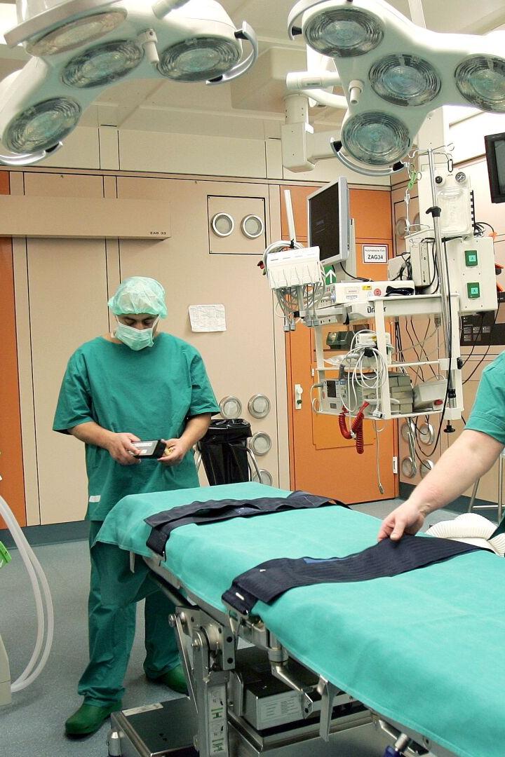 Zwei Ärzte in grüner OP-Kleidung bereiten einen Operationstisch in einem hell erleuchteten Operationssaal vor.