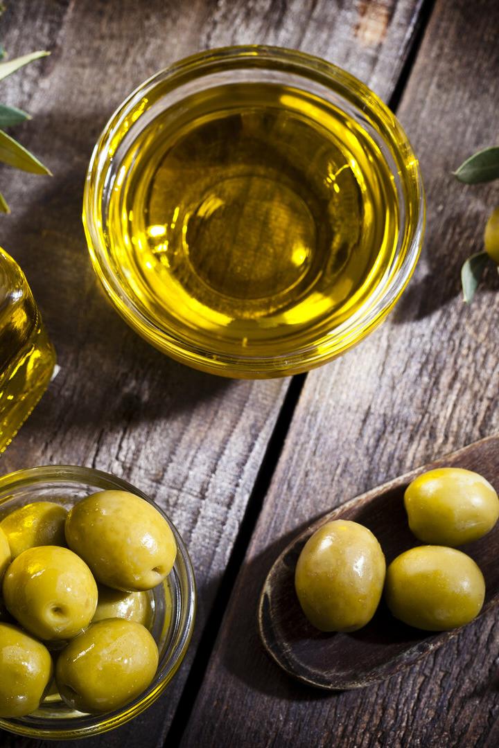 Grüne Oliven, Olivenöl in einer Schale und Flasche sowie Olivenzweige auf einem Holztisch.