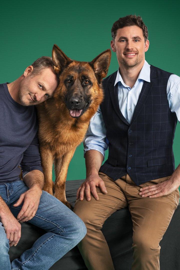 Zwei Männer posieren mit einem Deutschen Schäferhund vor einem grünen Hintergrund.