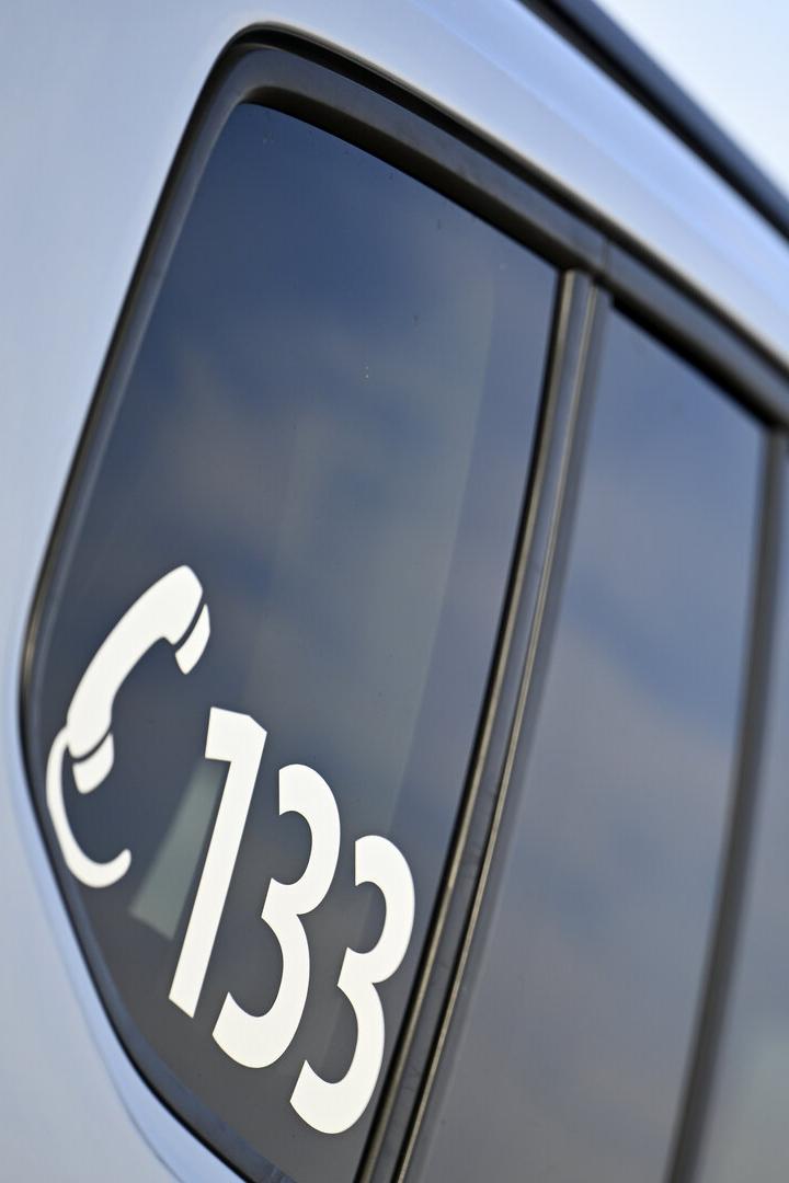 Auf der Autoscheibe ist in Weiß ein Telefonsymbol und die Nummer 133 zu sehen, im Hintergrund ein Blaulicht.