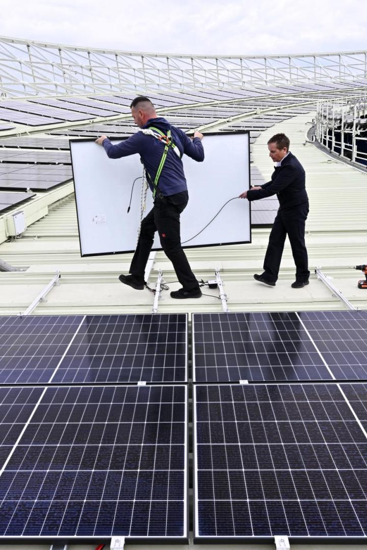 Zwei Männer installieren Solarpaneele auf dem Dach eines Stadions.