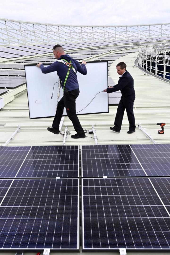 Zwei Männer installieren Solarpaneele auf dem Dach eines Stadions.