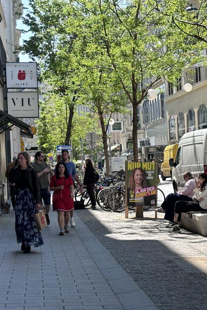 Auf einer belebten Straße sitzen zwei Frauen auf einer Bank, während Passanten vorbeigehen.