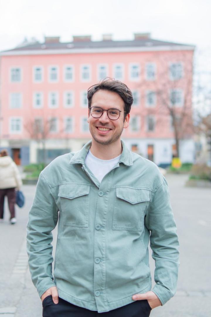 Ein lächelnder Mann mit Brille steht auf einem belebten Platz in der Stadt.