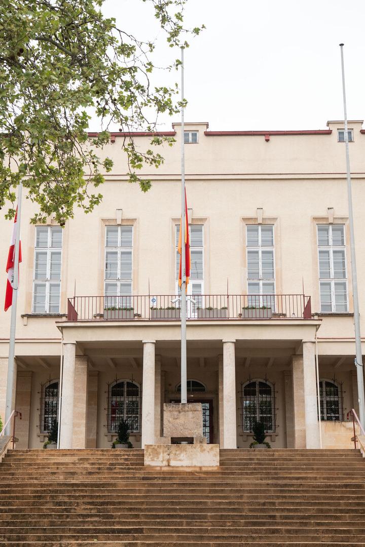 Beiges Verwaltungsgebäude mit Treppe, Säulen und Flaggen, von Bäumen eingerahmt.