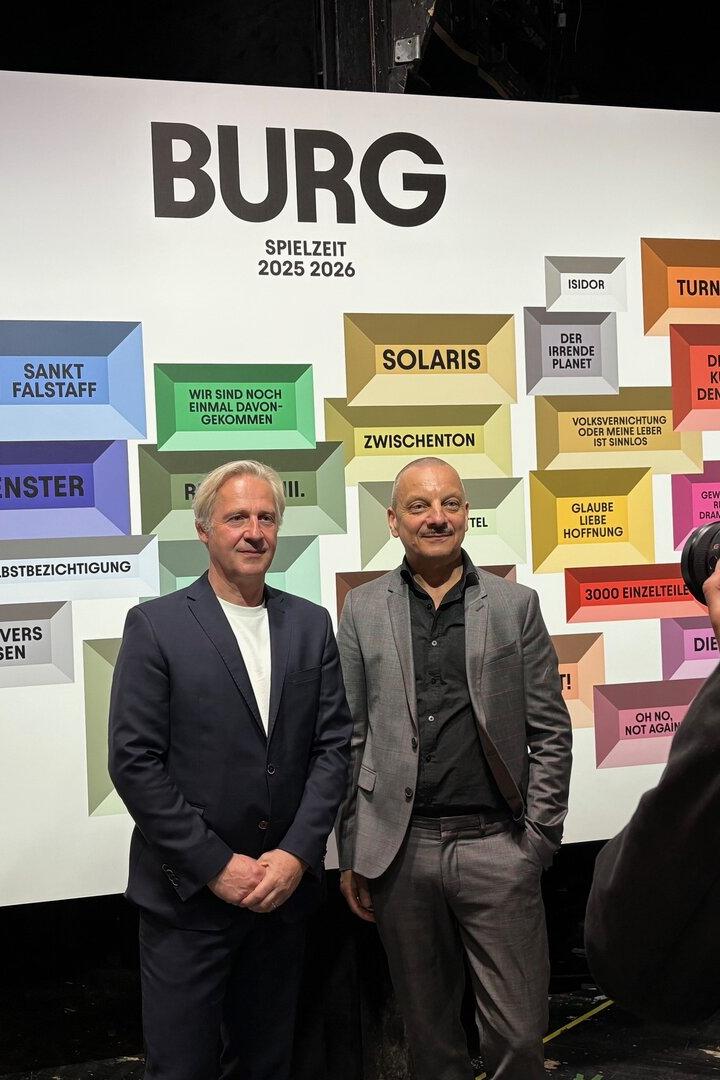 Zwei Männer posieren vor einer Wand mit dem Spielplan des Burgtheaters für die Saison 2025/2026.