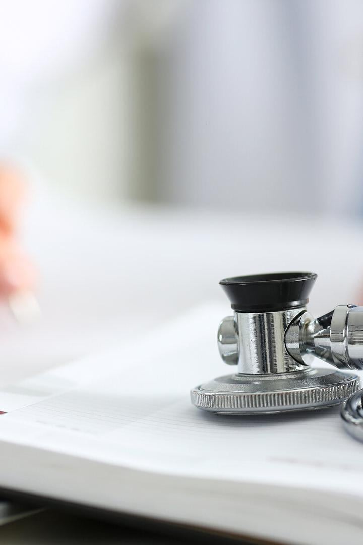 Ein Stethoskop liegt auf einem geöffneten Notizbuch, während eine Person im Hintergrund schreibt.