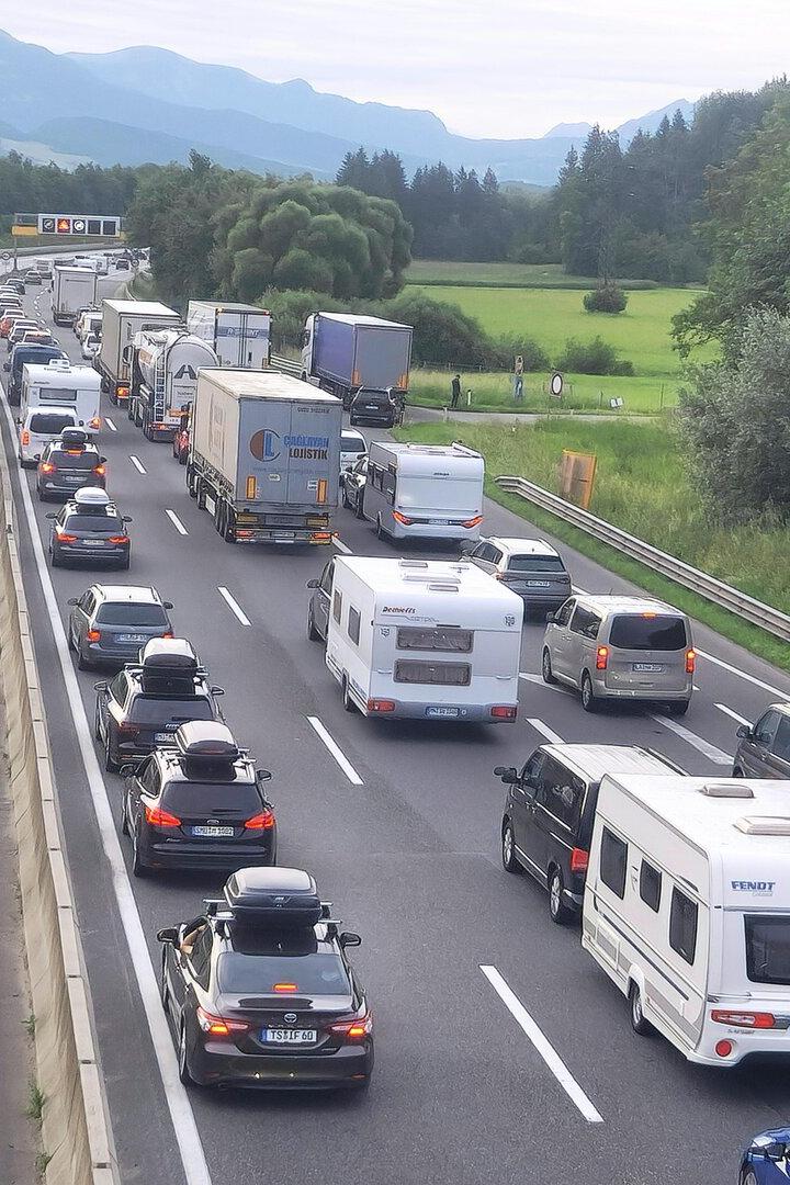 Stau auf der Autobahn mit vielen Autos und Lkw vor grüner Bergkulisse.
