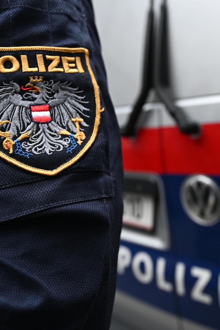 Ein Polizist mit österreichischem Wappen steht vor einem Einsatzfahrzeug mit rotem und blauem Streifen.