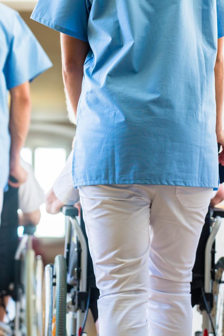 Zwei Pflegekräfte in blauer Kleidung schieben Rollstühle in einem hellen Flur.