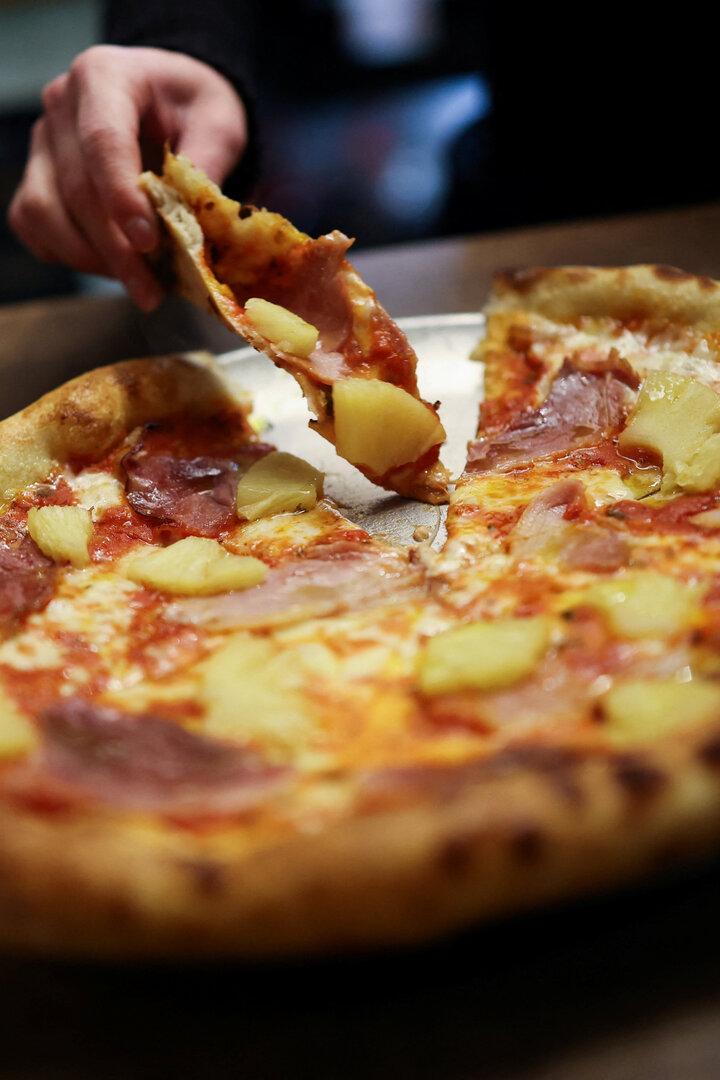 Eine Hand nimmt ein Stück Pizza mit Ananas und Schinken vom Teller.