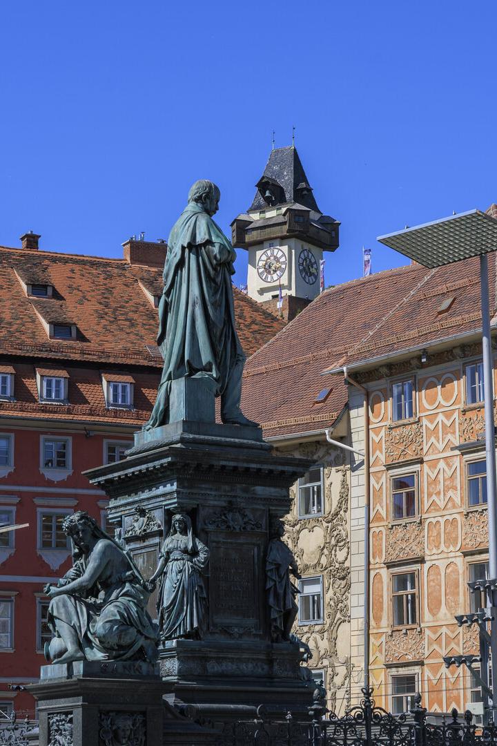 Eine Statue steht vor historischen Gebäuden in Graz, darunter der Grazer Uhrturm.