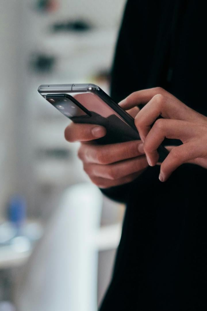 Eine Person tippt auf einem Smartphone, im Hintergrund ist ein unscharfer Arbeitsplatz mit Laptop zu sehen.