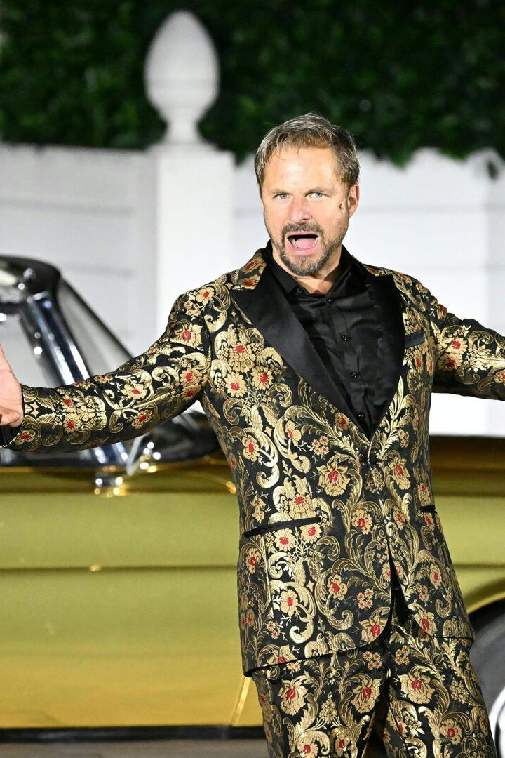 Ein Mann in einem auffälligen, gold-schwarz gemusterten Anzug gestikuliert vor einem goldenen Oldtimer mit Chauffeur.