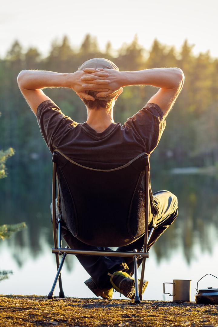 Man is sitting in a camping chair on the background of a forest lake on a beautiful summer evening.