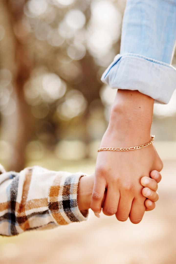 Eine Mutter und ihr Kind halten Händchen und gehen mit einem Schulrucksack durch den Park.