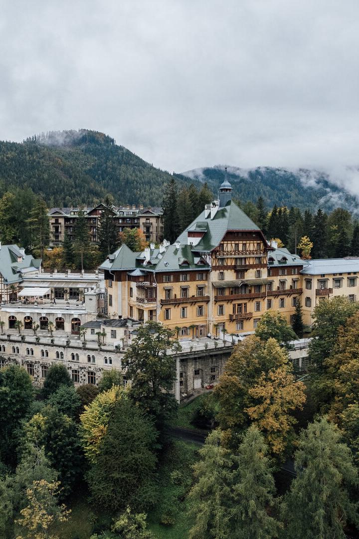 Das große Südbahnhotel mit seinen verschiedenen Trakten und großem Balkon. Es thront auf einem bewaldeten Hang. Im Hintergrund erheben sich Berge, Nebel lichtet sich