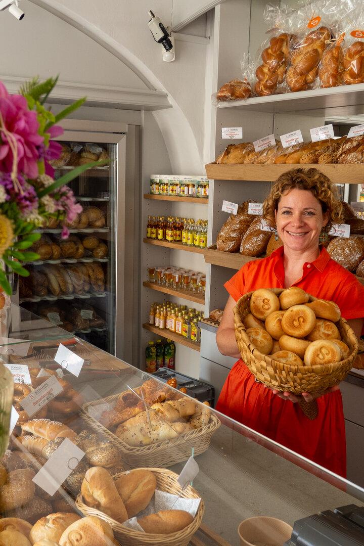 Eine Frau in rotem Kleid hält einen Korb mit Brötchen in einer Bäckerei, umgeben von Broten und einem Blumenstrauß.
