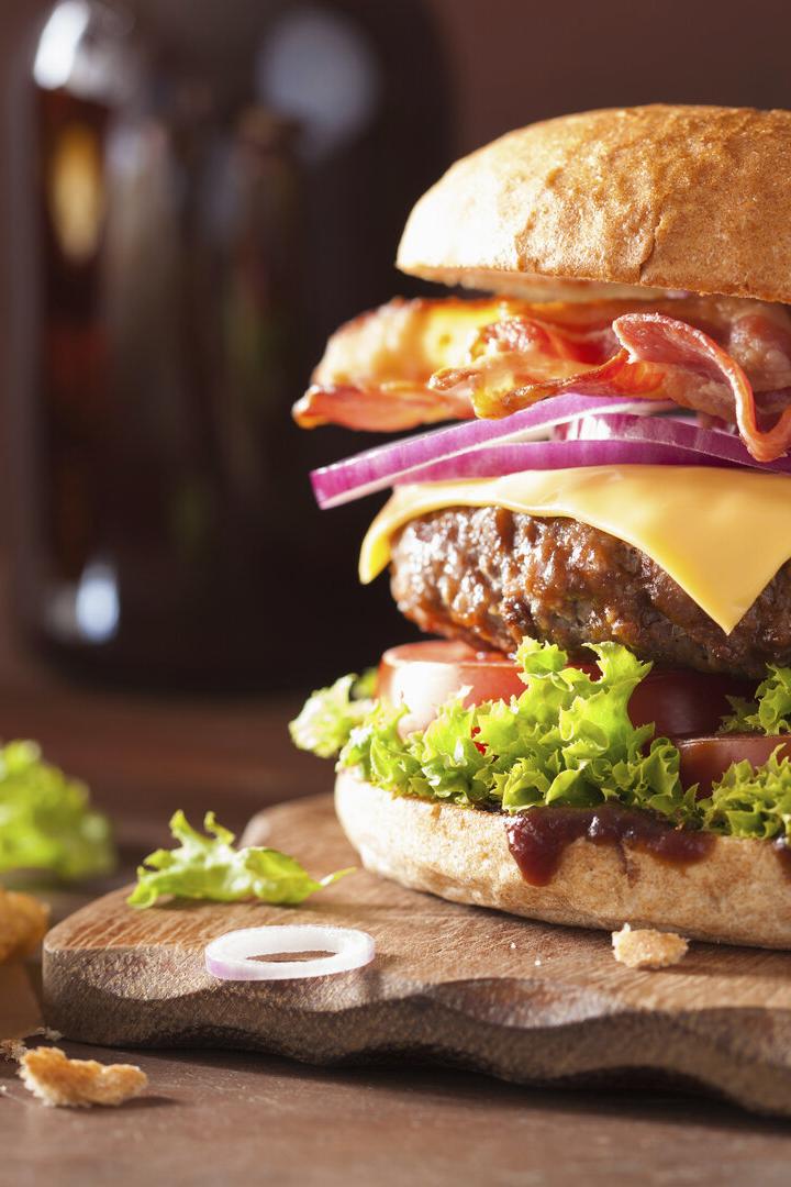 Ein Hamburger mit Speck, Käse, Tomaten und Salat, angerichtet auf einem Brett, daneben Pommes Frittes.