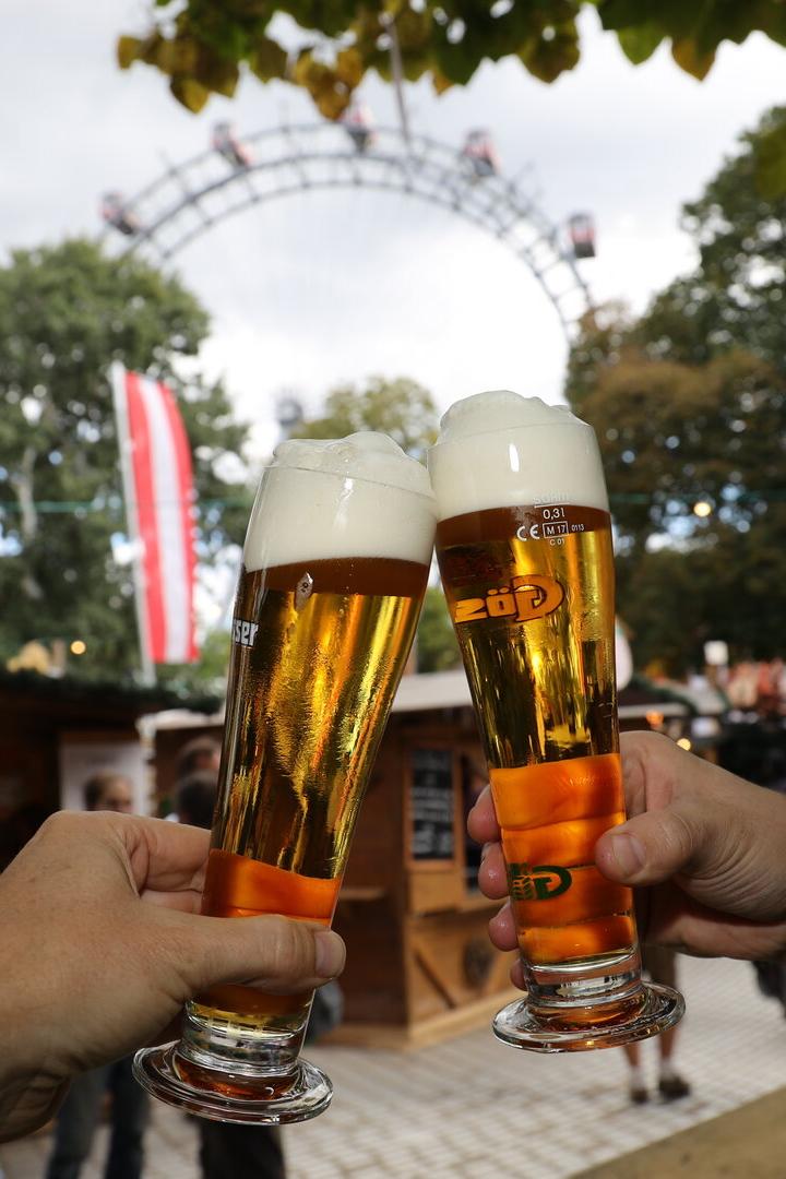 Zwei Hände mit Biergläsern, die gerade zusammenstoßen, im Hintergrund sieht man das Wiener Riesenrad