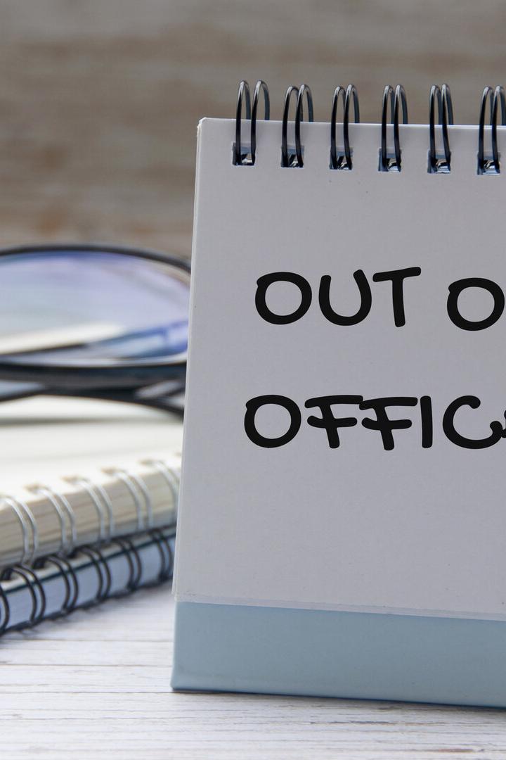 Ein Tischkalender zeigt die Aufschrift „OUT OF OFFICE“, daneben liegen eine Brille, Notizbücher und ein Stift.