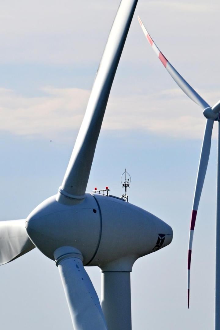 Zwei Windkraftanlagen mit rot markierten Rotorblättern vor einem hellen Himmel.