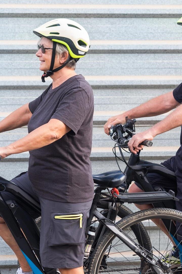 Happy senior couple doing together outdoor activity with electric bicycle. Healthy lifestyle. Yellow helmet, black clothes. Outdoor against a gray metal panel