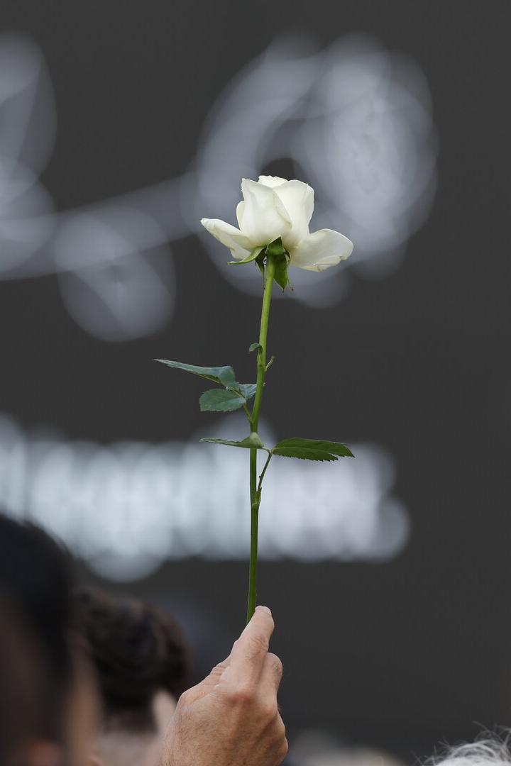 Eine Hand hält eine einzelne weiße Rose vor einem dunklen Hintergrund mit einer verschwommenen grafischen Darstellung einer Blume.