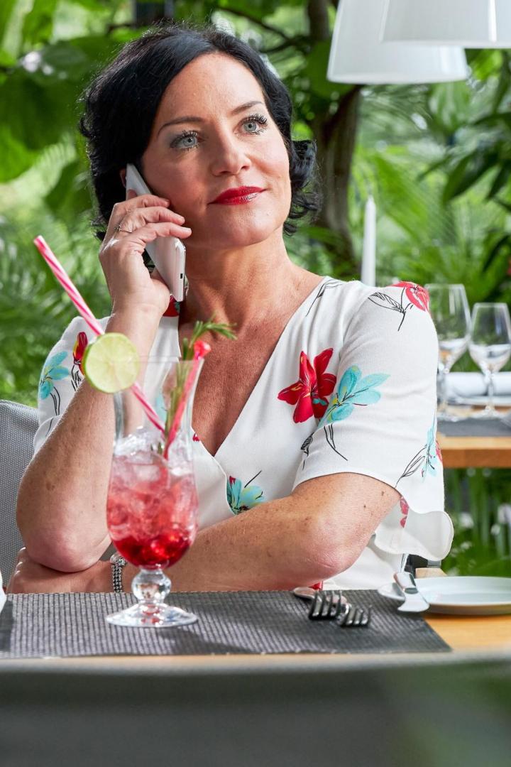 Eine Frau sitzt an einem gedeckten Tisch im Restaurant, telefoniert und hat einen Cocktail mit Limettenscheibe vor sich.