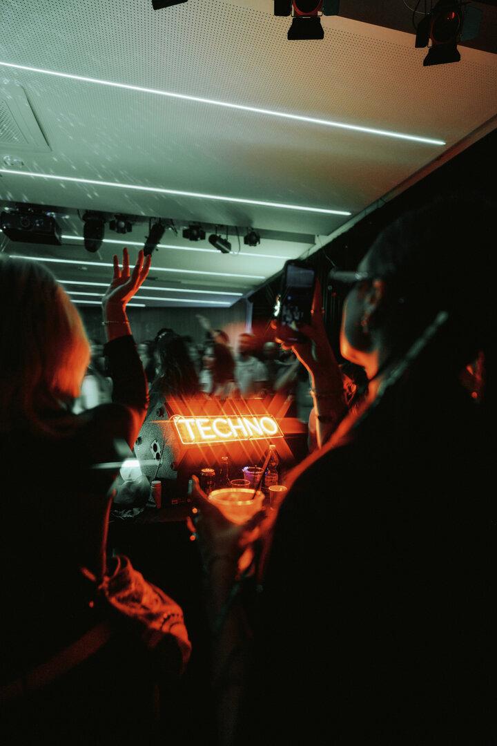 Blick vom DJ-Pult auf den Innenraum. Menschen haben die Hände in der Höhe und tanzen, eine Person fotografiert.. Auf einem Leuchtschild steht Techno.