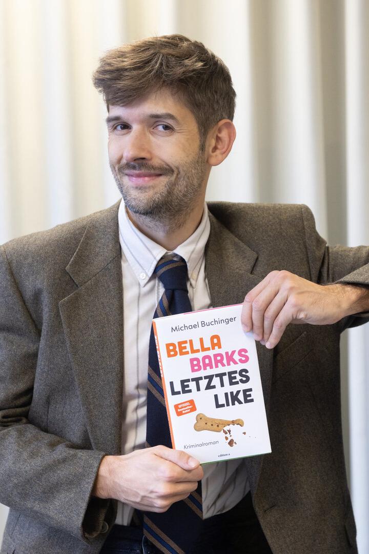 Ein Mann im Anzug hält ein Buch mit dem Titel „Bella Barks Letztes Like“ und lächelt leicht.