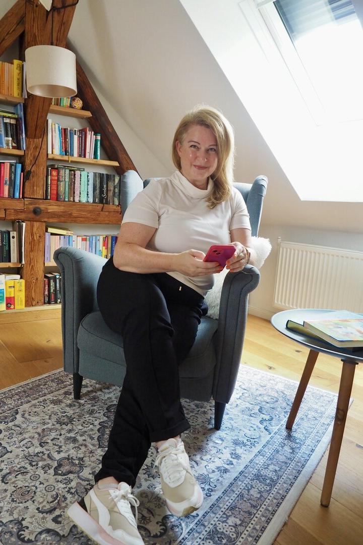Eine blonde Frau sitzt mit ihrem pinken Handy in einem grauen Ikeastuhl vor einer Bücherwand.