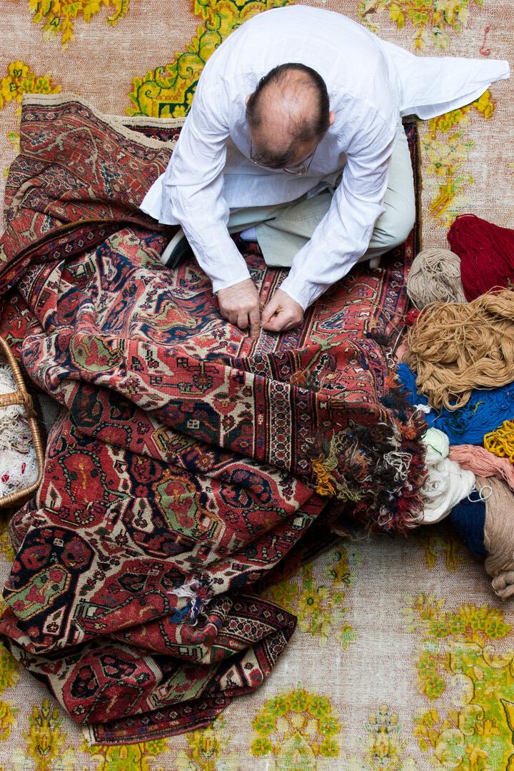 Vogelperspektive: Ein Mann hockt am Boden, vor ihm ein Teppich, neben ihm Wolle und Flickzeug. Er arbeitet am Perser mit klassischem Muster.