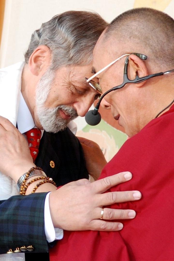 Nußbaumer und der Dalai Lama berühren respektvoll ihre Stirnen, einer trägt traditionelle rote Roben und ein Headset, der andere einen Anzug mit Schal.