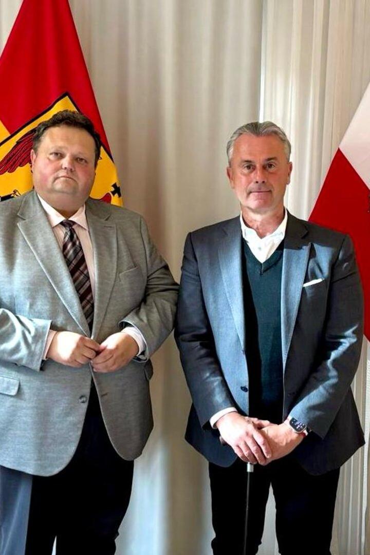 Zwei Männer im Anzug stehen nebeneinander vor einer rot-gelben und einer rot-weiß-roten Fahne mit Adler.