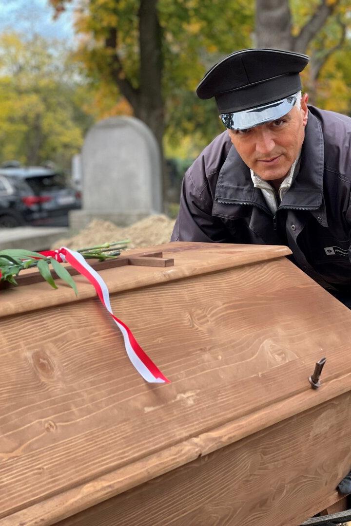 Ein Totengräber hebt einen Holzsarg mit Blumenschmuck in ein Grab.