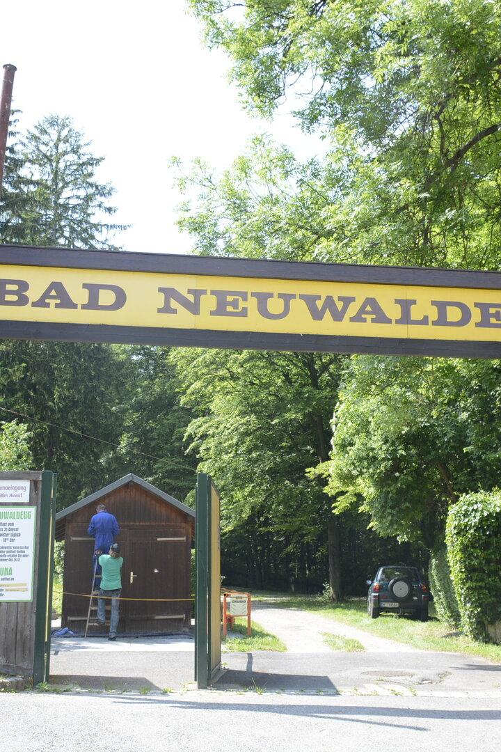 Ein Schild mit der Aufschrift "Bad Neuwaldegg" überspannt die Einfahrt, während zwei Personen an einem Häuschen arbeiten.
