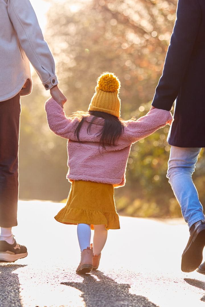 Ein Kind in gelber Mütze hält die Hände zweier Erwachsener und spaziert mit ihnen auf einem sonnigen Weg.