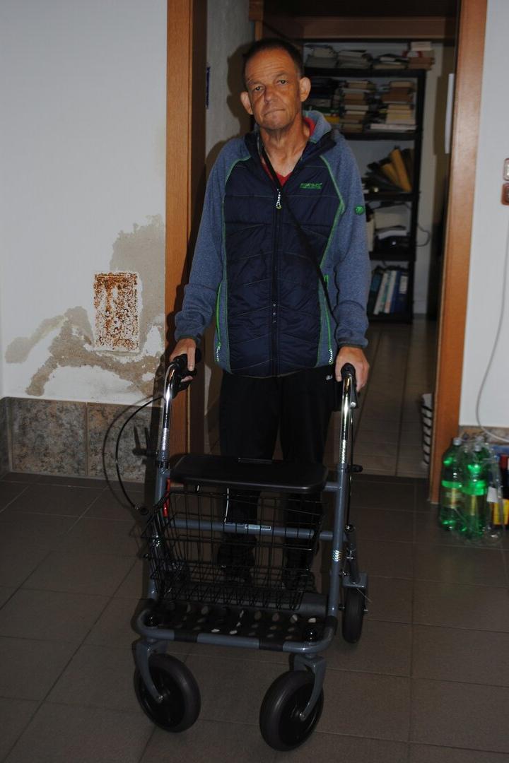 Ein Mann steht mit einem Rollator in einem Flur, im Hintergrund sind Regale mit Akten und Getränkeflaschen zu sehen.