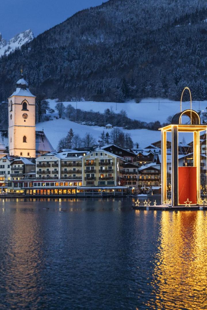 Beleuchtete Kirche und Häuser am verschneiten Seeufer des Wolfgangsees, große schwimmende Weihnachtslaterne auf dem Wasser, Berge im Hintergrund.