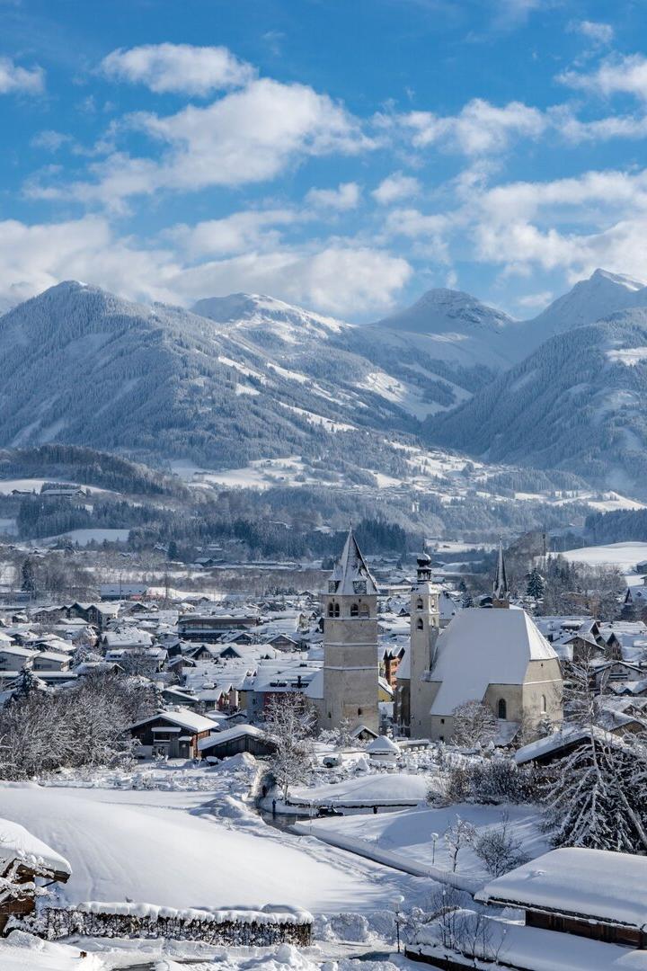 Blick von oben ins Tal auf Kitzbühel: Zwei Kirchen ragen heraus, dahinten erheben sich die Berge. Die Landschaft ist verschneit.
