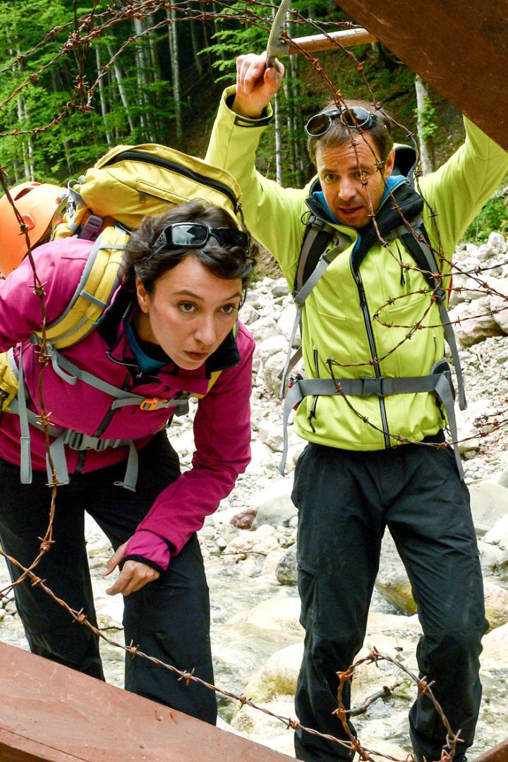 Felix Eitner an der Seite von Ursula Strauss mit Rucksäcken bücken sich vorsichtig unter Stacheldraht durch eine Metallkonstruktion im Gebirge.
