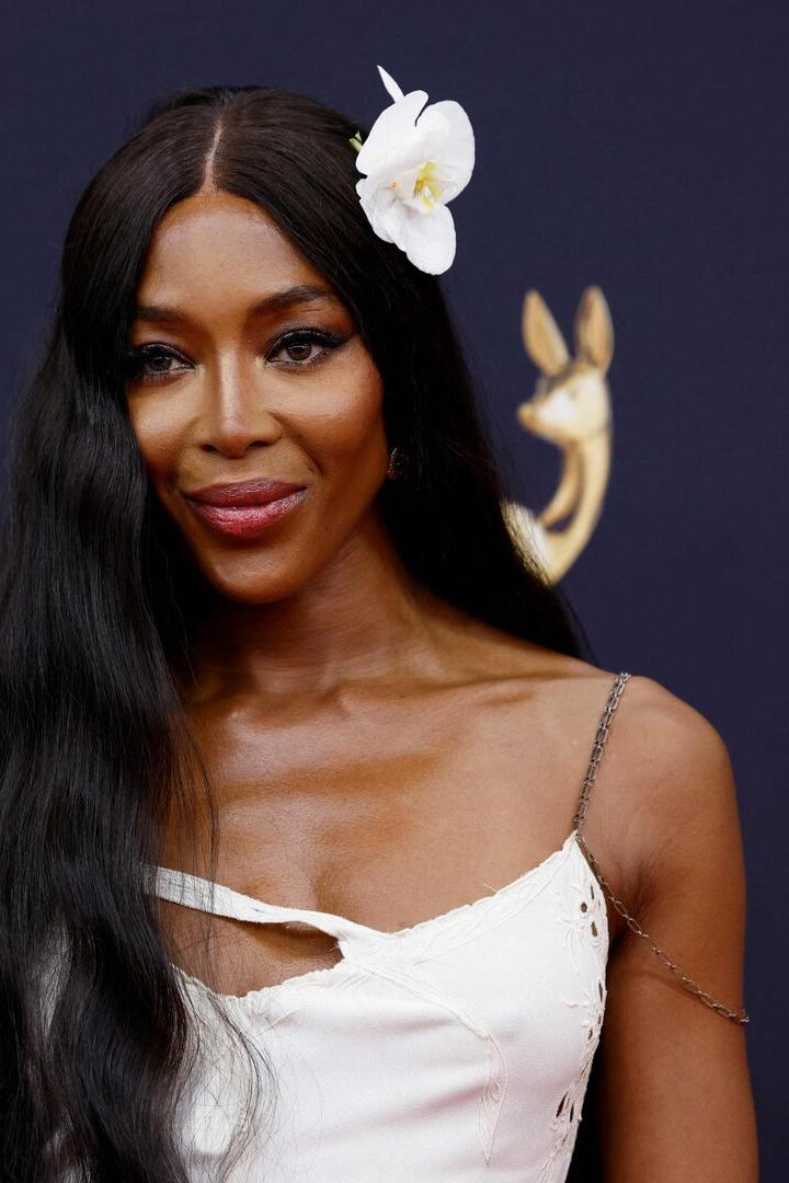 Naomi Campbell mit weißer Blüte im Haar und weißem Kleid vor dunklem Hintergrund.