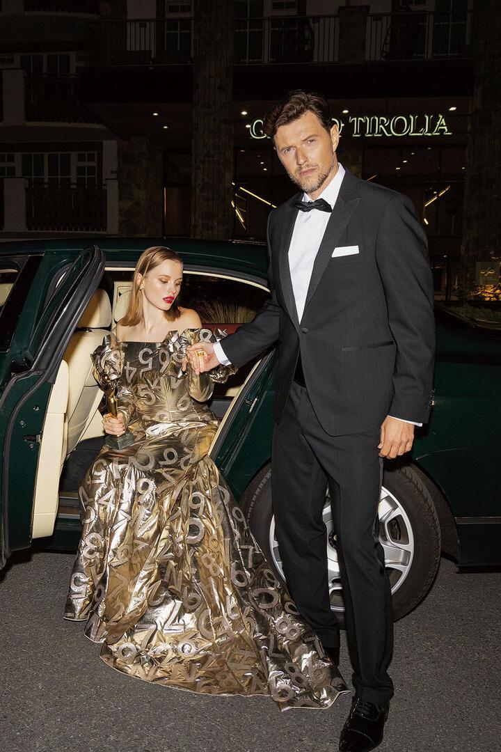 Ein Mann und eine Frau, die aus einem Auto steigen. Er trägt einen Smoking, sie ein goldenes Kleid. 