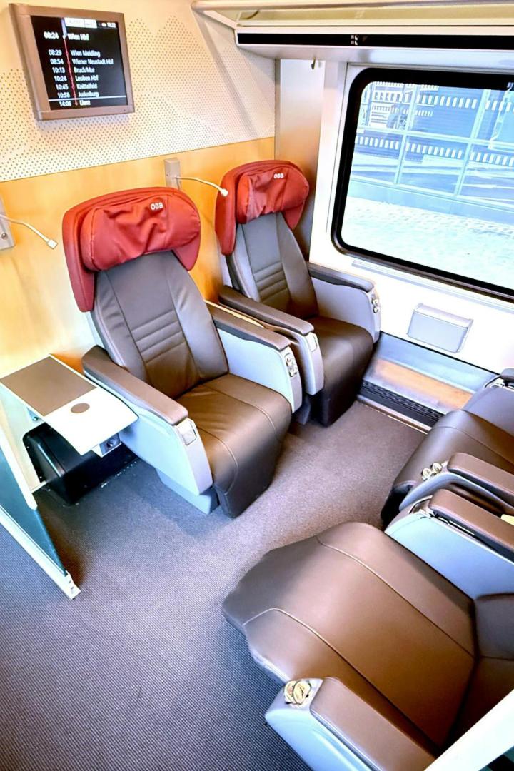 Vier bequeme Ledersitze mit roten Kopfstützen in einem modernen Zugabteil, großes Fenster und Monitor an der Wand.