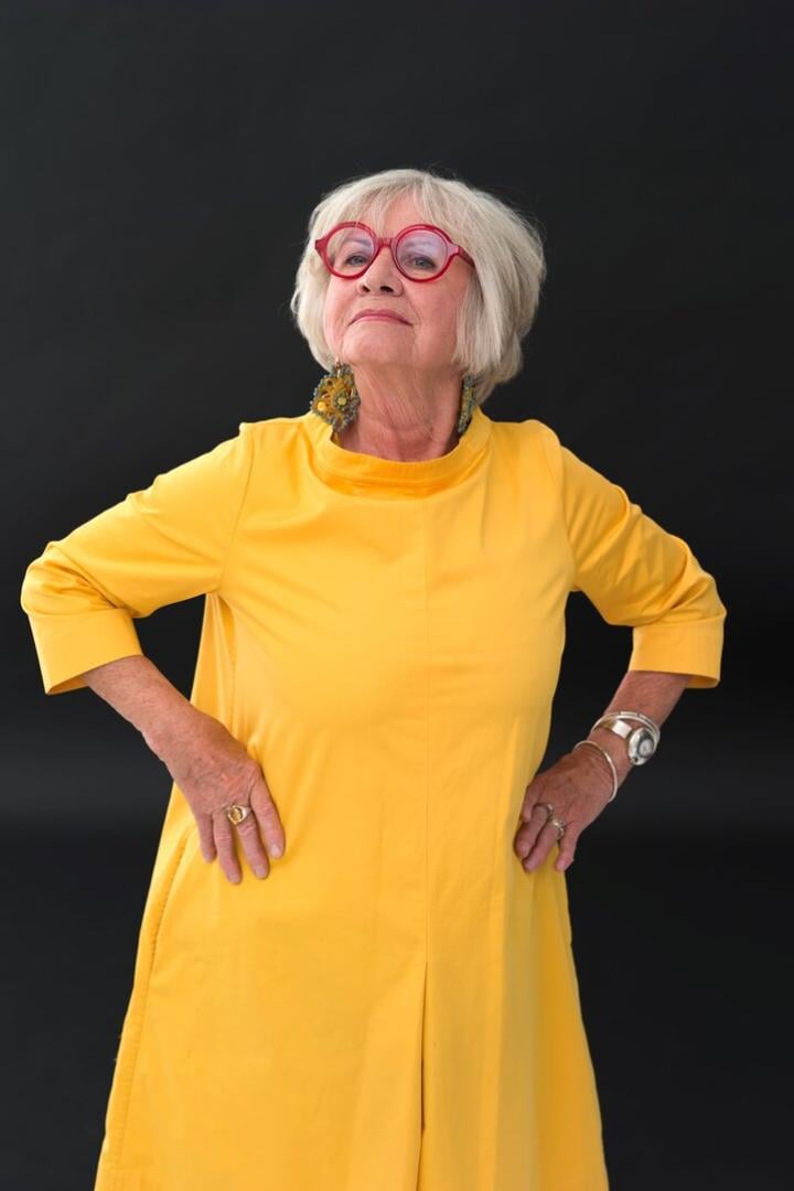 Frau im gelben Kleid und mit roter Hornbrille blickt in die Kamera