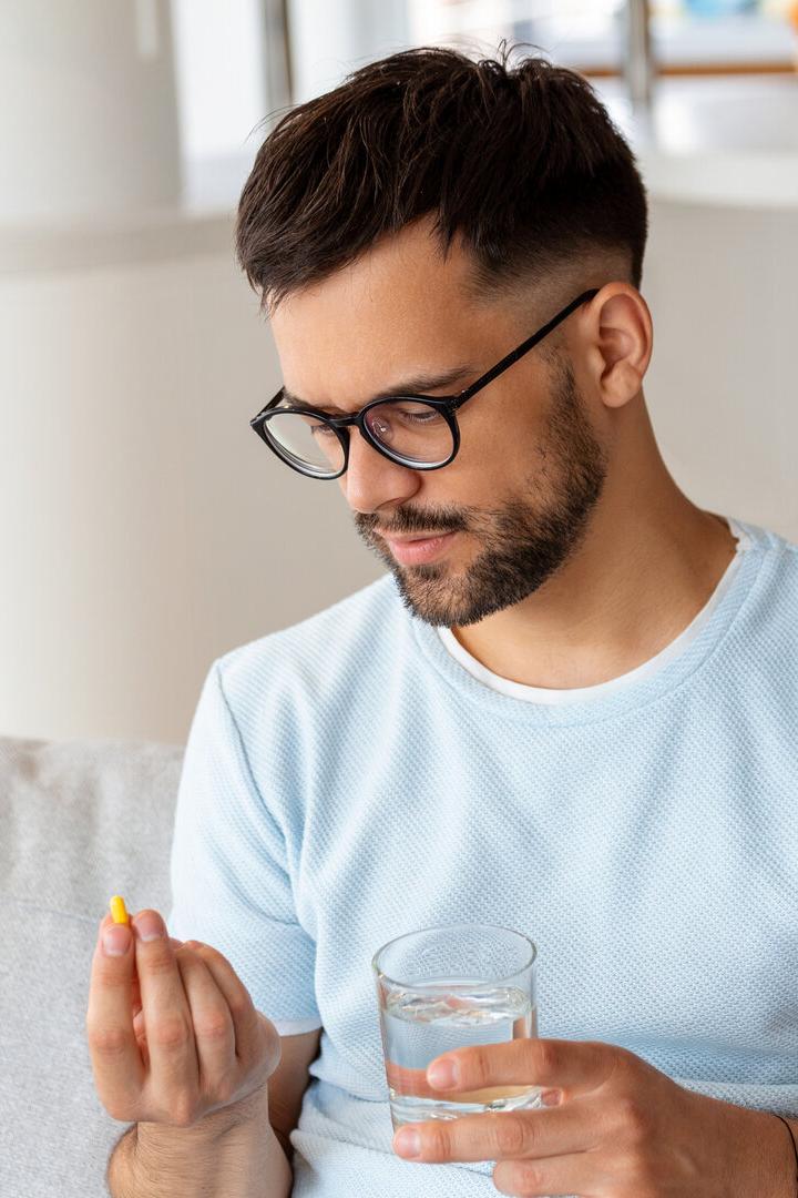 Ein Mann mit Brille hält ein Glas Wasser in der einen und eine gelbe Kapsel in der anderen Hand.