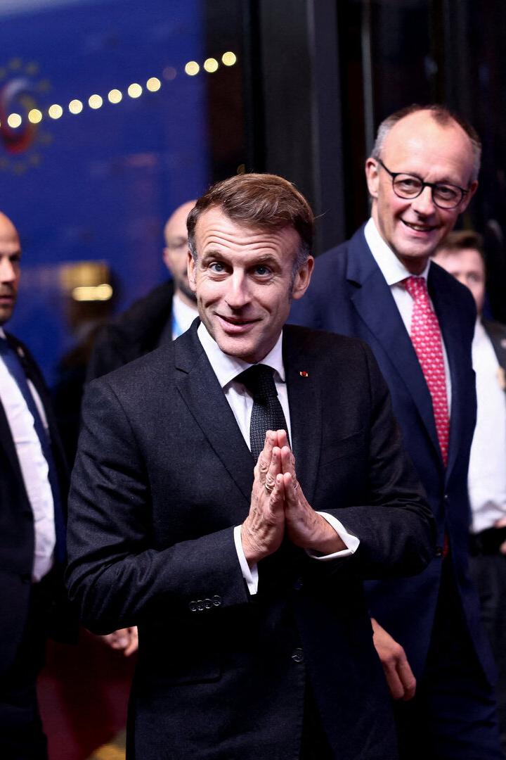 Emmanuel Macron mit gefalteten Händen, begleitet von Friedrich Merz und weiteren Personen.