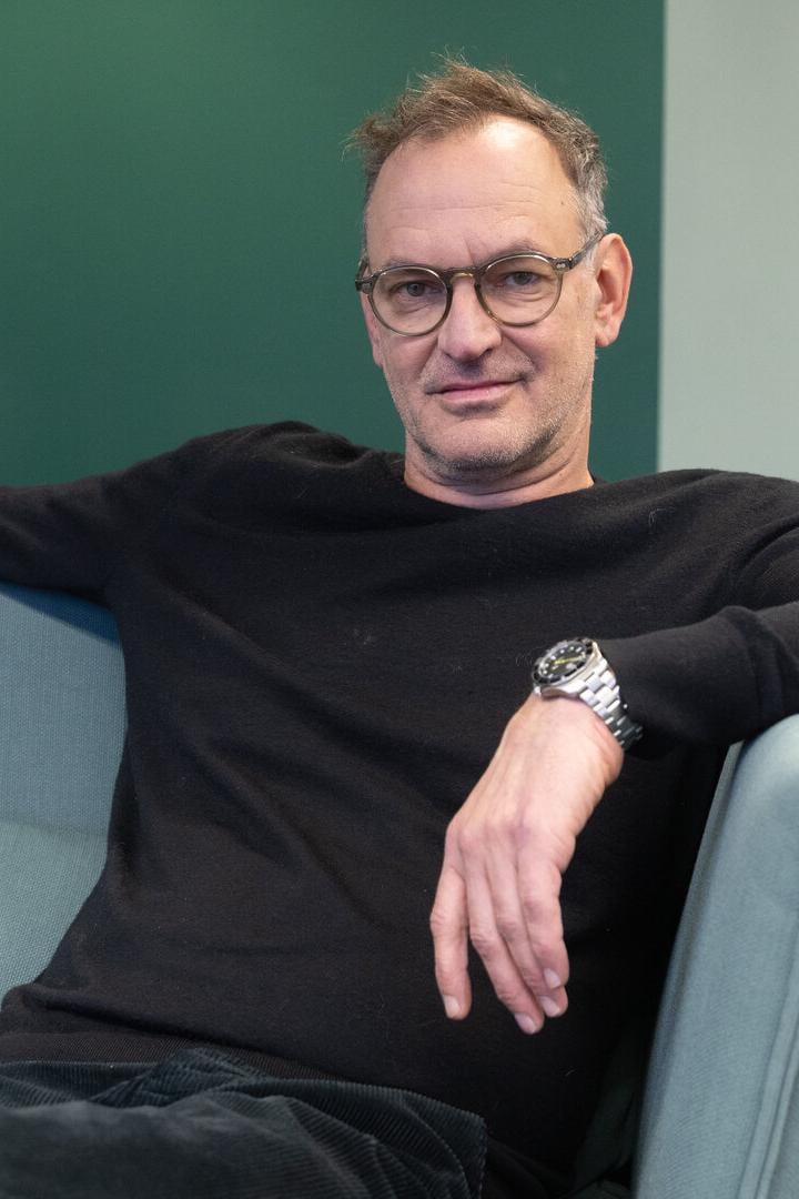 Ein Mann mit Brille und schwarzem Pullover sitzt entspannt auf einem hellgrünen Sofa vor einer grünen Wand.